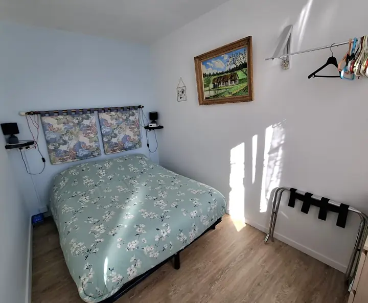 La chambre : le lit double et sa housse de couette à fleurs, sous un tableau de chaumière
