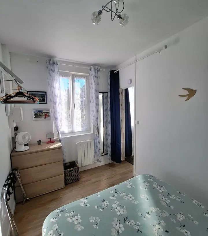La chambre vue de la porte : le lit, la fenêtre aux rideaux bleus et la commode